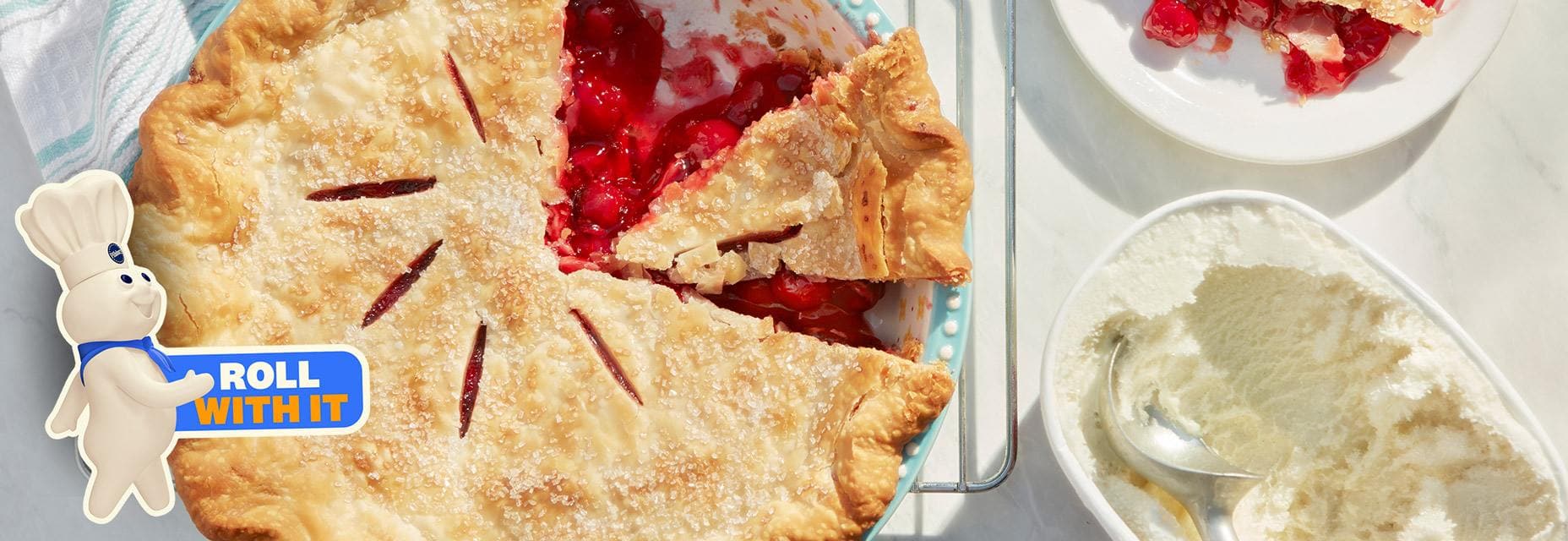 Cherry pie with a slice missing, alongside a plate with a slice and a bowl of whipped cream.