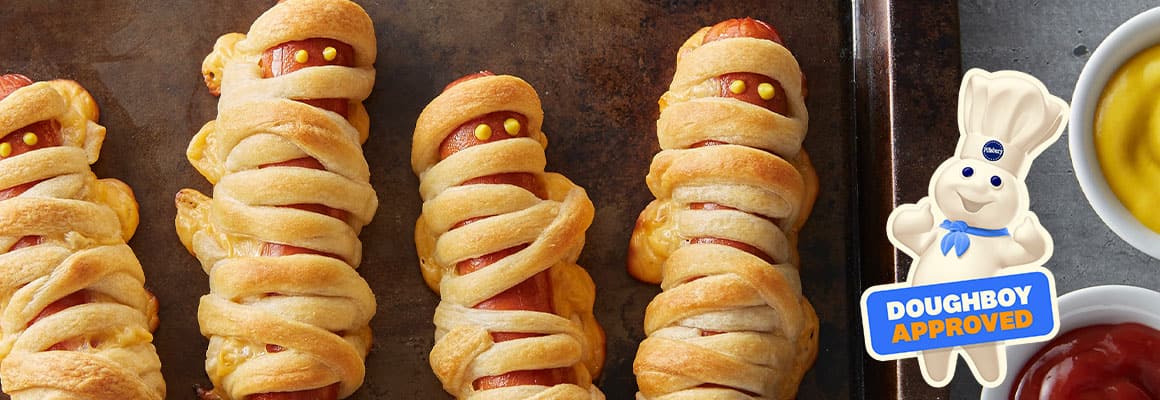 Five crescent mummy dogs on a baking tray with bowls of mustard and ketchup nearby. 